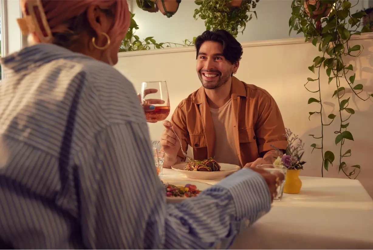 A smiling person holding a glass of drink, sitting at a table with another person, with green plants and a light-colored wall in the background.