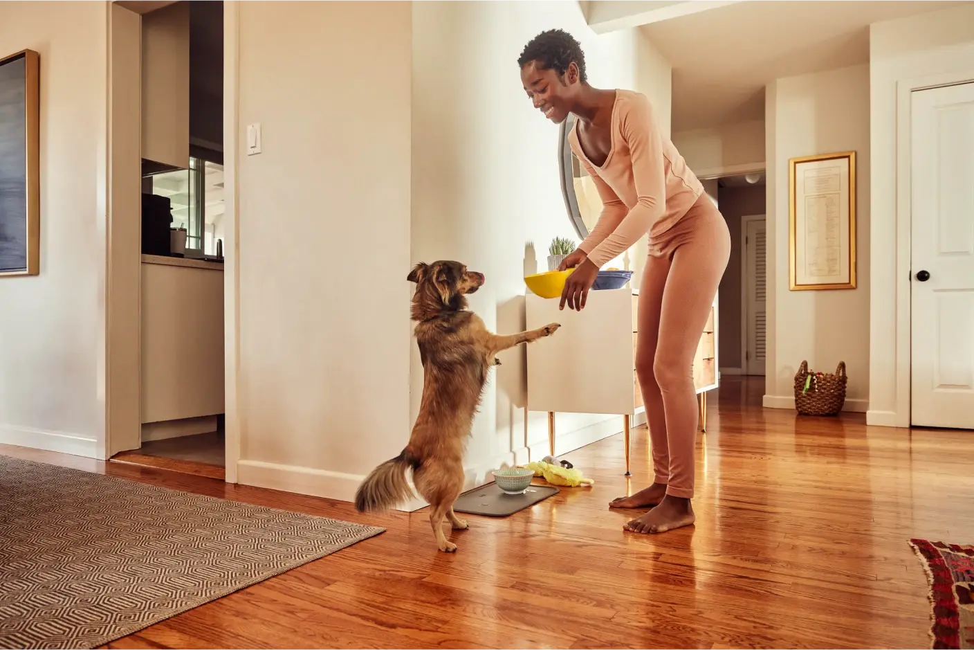 A woman in pink loungewear smiling and feeding her dog inside a modern, cozy home.